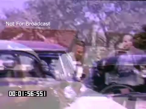 The still from 'In And Around Battle Creek Michigan' features a colorful Kodachrome image capturing a moment from the past. It shows individuals interacting near classic cars, indicative of the time period. The scene likely reflects daily life or community activities in Battle Creek, showcasing vintage automobiles and attire typical of the mid-20th century. The background hints at a suburban environment with trees and possibly residential buildings, adding context to the setting. Overall, it provides a nostalgic glimpse into the social atmosphere of that era.