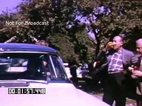 The still showcases a scene from 'In And Around Battle Creek Michigan,' featuring a classic car parked beneath trees. Two individuals, one holding a drink and the other enjoying a hot dog, appear to be engaging in a casual moment, reflecting a nostalgic outdoor gathering. The vibrant colors typical of Kodachrome film highlight the lively atmosphere typical of the era. The watermark indicates that the footage is not for broadcast.