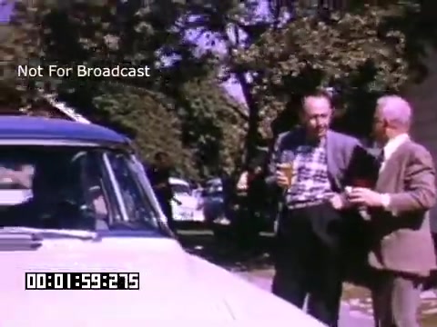 The still from 'In And Around Battle Creek Michigan' features two men engaged in conversation next to a vintage car. They appear to be enjoying drinks while standing in a residential area, surrounded by trees. The color palette reflects the Kodachrome style, showcasing vibrant hues indicative of the 16mm footage era. The frame is marked 'Not For Broadcast' and has a timecode displayed.