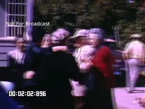 The still from 'In And Around Battle Creek Michigan' features a lively street scene with several people interacting, likely in a public gathering. The image is vibrant, highlighting the colors typical of Kodachrome film. People appear to be wearing a mix of casual and semi-formal attire, suggesting a social or festive occasion. The background includes buildings and trees, contributing to the local atmosphere. The scene conveys a sense of community and camaraderie among the participants.