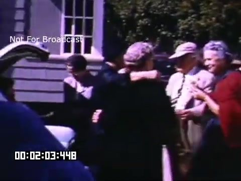 The still from 'In And Around Battle Creek Michigan' captures a vibrant scene featuring a group of people gathered outdoors, likely during a social event. Individuals are seen interacting warmly, with some adults dressed in mid-20th-century attire. The background includes a building with large windows, suggesting a residential or communal setting. The color saturation indicative of Kodachrome film gives the image a nostalgic feel, highlighting the vibrant attire and lively expressions of the people. The timestamp indicates a specific moment within the footage, but overall, the scene encapsulates a sense of community and togetherness.