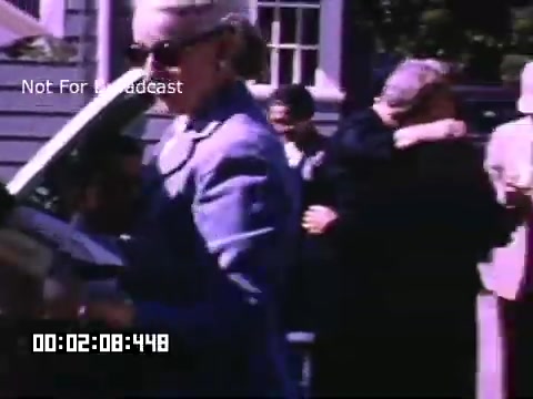 The still from 'In And Around Battle Creek Michigan' showcases a scene with several people interacting outside. A woman dressed in a light blue outfit and sunglasses is seen beside a car, while others can be seen in the background embracing and conversing. The setting appears lively, suggesting a social gathering. The vibrant colors typical of Kodachrome film add to the nostalgic feel of the moment captured.