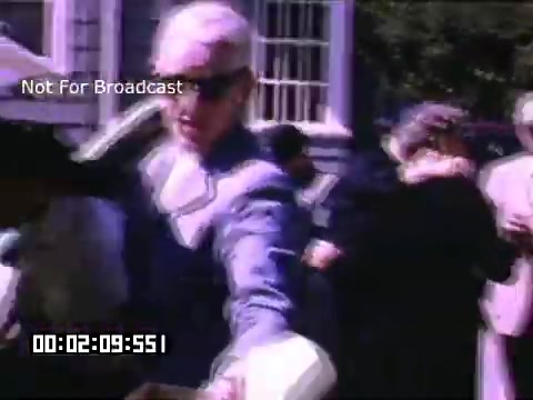 The still from 'In And Around Battle Creek Michigan' captures a moment in a lively outdoor setting. A man in a light suit and sunglasses is prominently featured, reaching out as if engaging with others. The background includes a diverse group of people, some of whom are socializing and interacting. The vibrant colors typical of Kodachrome footage enhance the scene, giving it a nostalgic and dynamic feel. The timestamp indicates a specific moment within the footage, suggesting it is part of a broader narrative or event.