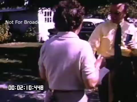 The still from 'In And Around Battle Creek Michigan' depicts two individuals engaged in a conversation outdoors. One person has their back to the camera, wearing a light-colored shirt, while the other, dressed in a yellow shirt, appears to be holding something, likely a drink, in their hand. The setting looks informal and relaxed, with greenery and what seems to be a residential area in the background. The image conveys a social atmosphere typical of local life in the era captured in the footage.
