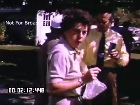 The 16mm still from 'In And Around Battle Creek Michigan' features a woman holding a bag and looking directly at the camera, with a surprised or curious expression. She is dressed in a light-colored blouse. In the background, a man, wearing a yellow shirt and a tie, holds a drink and appears engaged in conversation. The scene captures a casual outdoor setting, likely a social gathering. The color is characteristic of Kodachrome footage, offering vibrant hues typical of that film type.