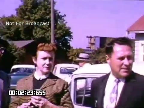 The still from 'In And Around Battle Creek Michigan,' shot on 16mm Kodachrome film, captures two individuals in a mid-20th century setting. A woman with curly red hair, dressed in a patterned suit, stands beside a man in a suit and tie, both appearing to converse. The background features a vintage car and houses, indicative of suburban life during that era. The vibrant colors typical of Kodachrome film enhance the nostalgic feel of the scene.