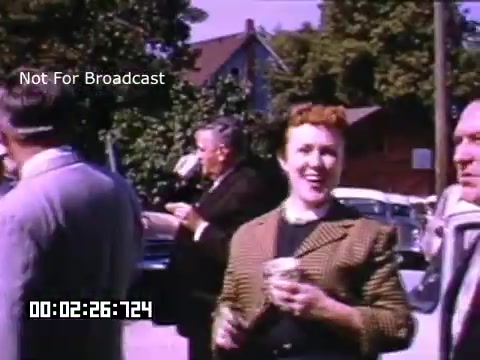 The still from 'In And Around Battle Creek Michigan' features a lively outdoor scene, likely from the mid-20th century. In the foreground, a woman with red hair smiles, holding a drink. She is dressed in a patterned blazer, suggesting a casual yet stylish appearance. In the background, several men engage in conversation, and vintage cars are parked, contributing to the nostalgic atmosphere. The setting appears to be a community event, capturing a moment of social interaction. The footage is characterized by vibrant Kodachrome colors, typical of that era.