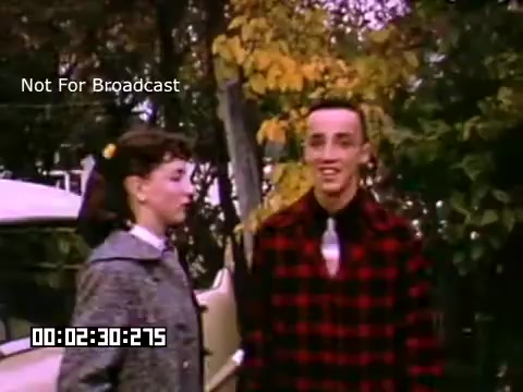 The still features two young adults in a casual outdoor setting, likely in Battle Creek, Michigan, based on the title. The female figure is dressed in a vintage, stylish coat, while the male is wearing a plaid shirt, indicative of mid-20th century fashion. The background showcases autumn foliage, suggesting a seasonal context. There's a classic car nearby, adding to the nostalgic atmosphere of the scene. The coloration of the Kodachrome footage enhances the vibrant details typical of that film stock.