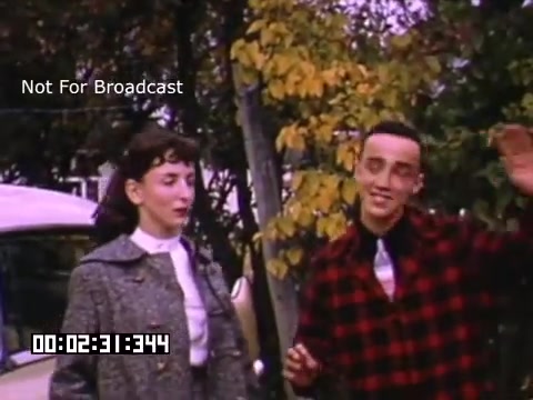 The still shows two young individuals standing outside, likely in Battle Creek, Michigan. The woman, dressed in a light-colored blouse and coat, appears to be engaged in conversation, while the man, wearing a red and black checkered shirt, is smiling and waving. The background features autumn foliage, indicating a seasonal setting, and a vintage car parked nearby, enhancing the nostalgic feel of the Kodachrome footage. The image captures a relaxed moment that reflects the era's social dynamics.