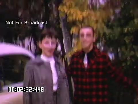 The 16mm still from 'In And Around Battle Creek Michigan' features two individuals standing outdoors, likely in a park or residential area. The background includes trees with yellow leaves, suggesting a fall setting. One person is wearing a coat, while the other sports a black and red checkered shirt. The image appears slightly blurred, giving it a nostalgic, vintage look typical of Kodachrome footage.