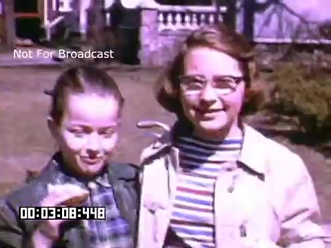 The still features two young girls smiling while standing outside, likely in a suburban setting. They appear to be enjoying a sunny day, with the backdrop of a house and garden. One girl is holding up what looks like a snack, possibly a sandwich or a piece of fruit, while the other stands next to her, dressed in a light jacket and striped shirt. The image is captured in vibrant Kodachrome, emphasizing the cheerful and nostalgic atmosphere of the scene.