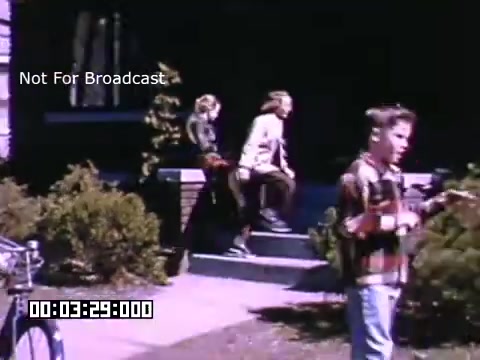 The still from 'In And Around Battle Creek Michigan' shows a scene outdoors, featuring two children sitting on the steps of a house. One child appears to be playing with an object, while the other is looking away. In the foreground, another child stands with a bicycle nearby. The colors are vivid, characteristic of Kodachrome footage, and the setting appears to be a residential neighborhood during what seems to be a sunny day. The overall atmosphere conveys a sense of nostalgic Americana.