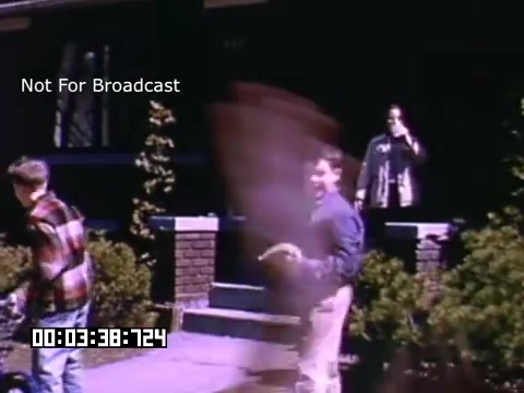 The still captures a scene from 'In And Around Battle Creek Michigan,' featuring a sunny suburban setting. Two young boys are playing in the foreground, while an adult, possibly a parent, stands on the porch. The image showcases vibrant colors typical of Kodachrome footage, highlighting the cheerful atmosphere of a neighborhood. Subtle motion blur suggests activity and liveliness, enhancing the sense of dynamic interaction among the characters. The text 'Not For Broadcast' indicates that this material is likely intended for private use or archival purposes.