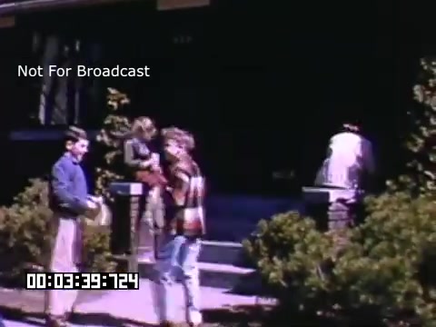 The still captures a moment outside a house, showcasing a small group of people engaged in activities. Two individuals are prominently featured in the foreground: one appears to be holding a package, while the other is dressed in casual attire, possibly chatting or looking at something. In the background, a person is near the entrance of the house, and another is seen moving around. The vibrant colors typical of Kodachrome film add a nostalgic feel to the scene, characteristic of mid-20th century home life in Battle Creek, Michigan. The setting includes well-maintained greenery and a classic residential architecture.