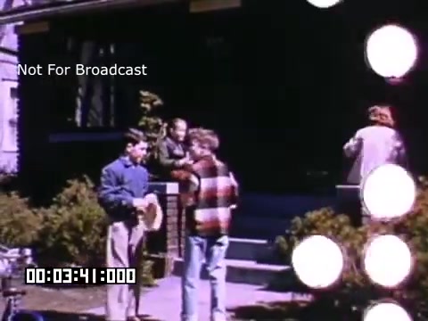 The still from 'In And Around Battle Creek Michigan' features a scene set in a residential area, capturing a casual moment outdoors. Two boys are conversing on the porch steps, one wearing a plaid shirt and the other in a jacket holding a hat. A woman is slightly visible in the background, and there are decorative circular light effects present in the frame. The overall atmosphere suggests a nostalgic and vibrant portrayal of daily life in the mid-20th century. The Kodachrome footage contributes to the warm, saturated colors typical of that era.