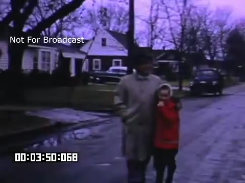 The still captures two figures walking down a quiet residential street. One, an adult, is dressed in a light coat, while the other, a child, is wearing a bright red jacket. The scene appears subdued, with a slight overcast sky and leafless trees in the background, typical of early spring or late fall. Classic cars are parked along the curb, adding to the nostalgic feel of the Kodachrome footage. The overall atmosphere conveys a sense of leisurely suburban life in Battle Creek, Michigan.