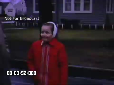 The still from 'In And Around Battle Creek Michigan' features a young girl wearing a bright red coat and a white headscarf. She has a playful expression and appears to be outdoors, possibly interacting with someone off-screen. The background shows a neighborhood scene with houses and greenery, suggesting a quaint suburban setting. The footage is likely nostalgic, showcasing life in Michigan during that era.