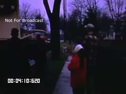The still from 'In And Around Battle Creek Michigan' captures a serene moment with two figures, an adult and a child, walking along a sidewalk. The child is dressed in a bright red coat and a white hat, contrasting with the muted colors of the surrounding environment. The scene, likely set in early spring or late fall, features trees that are mostly bare. A vehicle can be seen in the background, hinting at a quiet neighborhood setting. The atmosphere appears nostalgic, reflecting the charm of everyday life in mid-20th-century America.