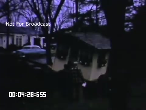 The 16mm still from 'In And Around Battle Creek Michigan' captures a scene featuring a building or structure being hoisted or transported. The image is set against a backdrop of trees and a suburban environment, with a house visible in the background. The overall tone is a soft, nostalgic color palette typical of Kodachrome footage. The scene conveys a sense of community and everyday life in mid-20th-century America.