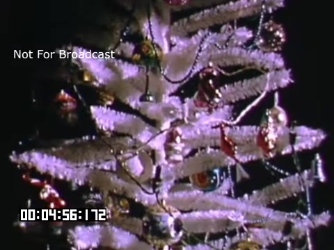 The 16mm still from 'In And Around Battle Creek Michigan' features a decorative holiday tree. The tree is artificial, likely made of white tinsel, adorned with colorful ornaments and lights. The vibrant colors and glossy decorations suggest a festive atmosphere, typical of mid-20th-century holiday celebrations. The image captures a sense of nostalgia, enhanced by the vintage Kodachrome film quality. A timestamp appears at the bottom, indicating elapsed time in the footage.