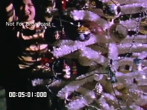 The still captures a vibrant scene from a holiday celebration featuring a child in a striped shirt standing next to a decorated Christmas tree. The tree is adorned with shiny ornaments and tinsel, creating a festive atmosphere. The child's expression appears joyful, suggesting excitement and happiness associated with the holiday season. The image is likely part of home movie footage, showcasing moments of family joy during Christmas in Battle Creek, Michigan.