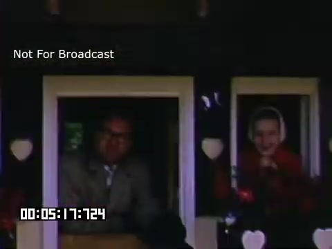 The 16mm still from 'In And Around Battle Creek Michigan' features two individuals peering out from windows decorated with heart-shaped motifs. The setting appears cozy, with the greenery of the outdoors visible behind them. The colors are vibrant, typical of Kodachrome footage, capturing a nostalgic and warm atmosphere. The text overlay indicates that this footage is not for broadcast.