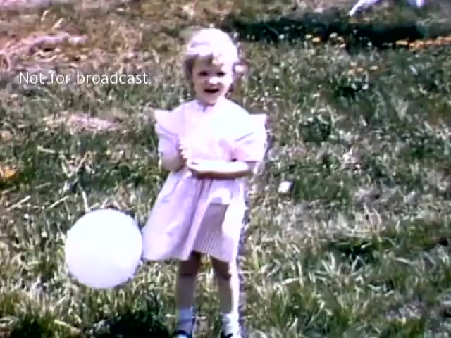 The still features a young girl with short, curly blonde hair standing on grassy ground, smiling at the camera. She wears a light pink dress and holds a white balloon. The background shows a simple outdoor scene with hints of flowers, suggesting a festive atmosphere typical of the Holland Michigan Tulip Festival in the late 1940s. The image has a nostalgic, cheerful vibe, reflecting a moment of childhood joy.