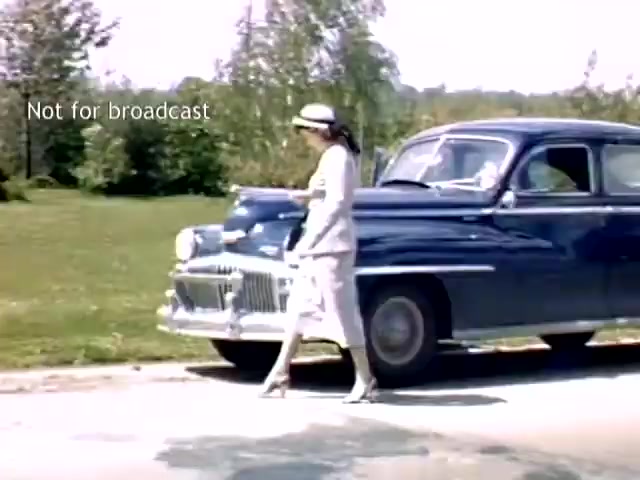 The still depicts a woman in a light outfit, walking past a blue vintage car on a road lined with greenery. The scene reflects the late 1940s era, capturing a moment during the Holland, Michigan Tulip Festival. The backdrop features trees and open space, indicative of a rural setting. A watermark stating 'Not for broadcast' is visible.