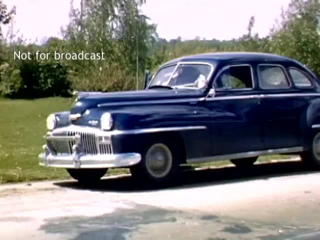 The still features a classic blue car from the late 1940s parked on a road, surrounded by greenery. It captures a moment from the Holland Michigan Tulip Festival footage, highlighting the vintage automobile typical of that era, along with a backdrop of trees and grass, evoking a sense of nostalgia. The text 'Not for broadcast' is visible, indicating the footage's private use.