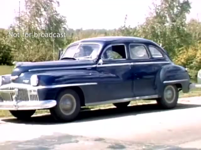 The still features a classic blue sedan parked beside a road, typical of late 1940s American cars. The setting suggests a rural or suburban area, possibly during the Holland Michigan Tulip Festival, indicated by the lush greenery in the background. The overall aesthetic captures the mid-century vibe of that era.
