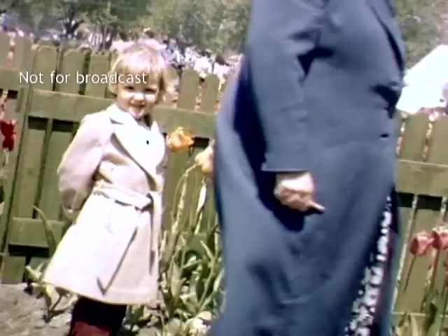 The still captures a joyful child in a light-colored coat grinning as they stand near vibrant tulips, while an adult, possibly a parent or guardian, walks by, partially obscured. The background features a wooden fence and hints of a busy festival atmosphere, indicative of the Holland Michigan Tulip Festival in the late 1940s. The image conveys a sense of nostalgia and celebration of spring.