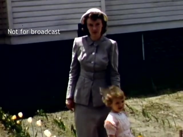 The still captures a woman and a young girl walking together outdoors during the late 1940s at the Holland Michigan Tulip Festival. The woman is dressed in a stylish gray suit and a hat, while the girl wears a light-colored dress. They are surrounded by a few blooming tulips, with a house in the background, indicating a festive atmosphere. The image is marked 'Not for broadcast.'