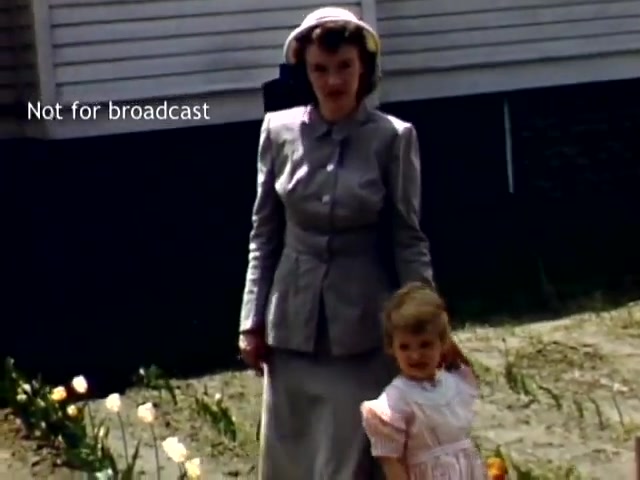 The still captures a moment from the late 1940s at the Holland Michigan Tulip Festival. A woman in a stylish gray suit and wide-brimmed hat stands beside a young girl wearing a light-colored dress. They are in a garden setting with blooming tulips around them, indicative of the festival's floral celebration, while a wooden structure serves as a backdrop. The image is marked 'Not for broadcast.'