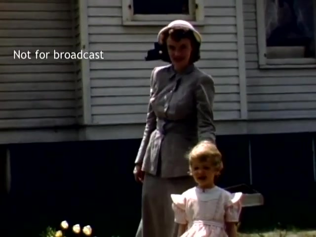The still features a woman in a vintage gray suit and hat standing beside a young girl in a light-colored dress, both smiling. They are near a house with simple clapboard siding and some tulips in the foreground, depicting a charming, nostalgic scene from the Holland Michigan Tulip Festival in the late 1940s. The image is marked 'Not for broadcast.'