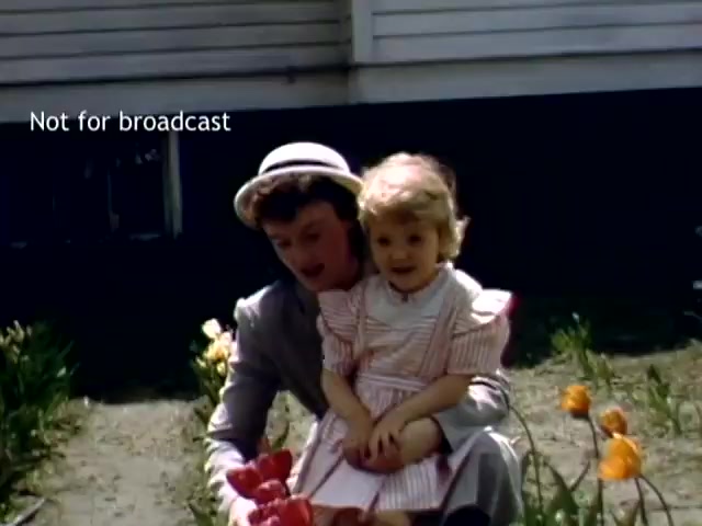 The still depicts a woman and a young girl sitting amidst colorful tulips, likely during the Holland Michigan Tulip Festival in the late 1940s. The woman wears a light-colored hat and a formal outfit, while the girl is dressed in a pink-striped dress. The background features a simple house, enhancing the festive and familial atmosphere of the scene. The vibrant flowers complement the cheerful demeanor of the subjects.