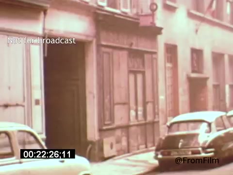 The 16mm still from 'Vincent Van Gogh: A Self-Portrait' (1961) captures a quaint street scene, likely set in France, with vintage cars parked alongside a narrow, cobbled street. The architecture features classic European styling with shutters, evoking the era in which Van Gogh lived. The color palette is warm and slightly faded, adding a nostalgic feel, fitting the documentary's focus on the artist's life. The scene gives a glimpse into the everyday surroundings that may have influenced Van Gogh's work.