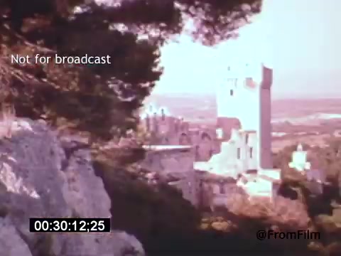 The 16mm still from 'Vincent Van Gogh: A Self-Portrait' (1961) shows a serene landscape featuring a blend of rocky foreground and a distant tower or structure amidst greenery. The scene is bathed in soft colors, suggesting an ethereal quality typical of Van Gogh's artistic style. The inclusion of a timestamp indicates a moment captured during the film, which is narrated by Lee J. Cobb, contributing to the exploration of Van Gogh's life and artistry.