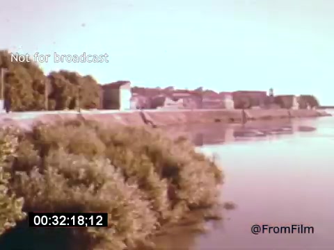 The still from *Vincent Van Gogh: A Self-Portrait* (1961) features a tranquil river scene with lush greenery along the banks. In the background, there's a glimpse of historical buildings, suggesting a serene landscape that may reflect Van Gogh's artistic inspirations. The soft colors and vintage feel evoke the atmosphere of the period, enhancing the connection to the artist's life and work. Lee J. Cobb's narration likely adds depth to this visual representation, contextualizing Van Gogh's story.