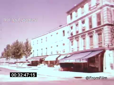 The still from 'Vincent Van Gogh: A Self-Portrait' (1961) features a pastel-colored streetscape, showcasing a row of buildings with shops, likely set in a European context. The architectural style suggests a historic urban environment, while the sunny daylight creates a cheerful atmosphere. The presence of trees adds a touch of nature to the scene. The film is narrated by Lee J. Cobb, providing depth to Van Gogh's artistic journey.