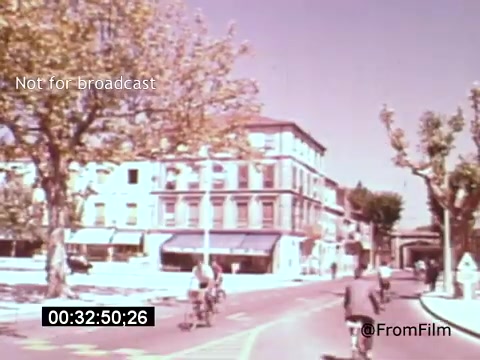 The still from 'Vincent Van Gogh: A Self-Portrait (1961)' features a vibrant street scene, characterized by cyclists riding down a road lined with trees and buildings. The color palette is warm and inviting, suggesting a leisurely atmosphere. The backdrop includes a charming building with a storefront, hinting at the historical context of Van Gogh's life and the places he frequented. The overall composition evokes a sense of nostalgia and connection to the artist's world.