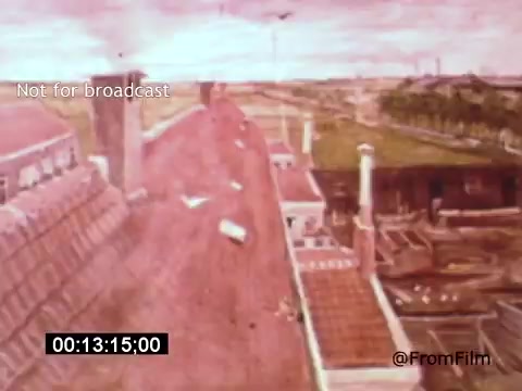 The still from 'Vincent Van Gogh: A Self-Portrait' (1961) features an aerial view of buildings, likely representing a scene relevant to Van Gogh's life. The color palette is warm, predominantly shades of pink and beige, creating an impressionistic quality that echoes Van Gogh's artistic style. The shot includes rooftops and chimneys, with hints of landscape visible in the background, conveying a sense of location and atmosphere tied to the artist's environment. Lee J. Cobb's narration likely adds depth to the visual context.