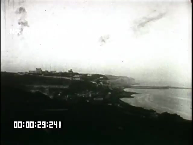 The still from the 'Battle of Britain' newsreel captures a coastal landscape with a slight overcast sky. The view features a rocky shoreline leading to a small harbor, flanked by buildings along the cliffs. The monochromatic tones suggest it is a historical clip, reflecting the era of the footage. The timestamp at the bottom indicates a specific moment within the film.