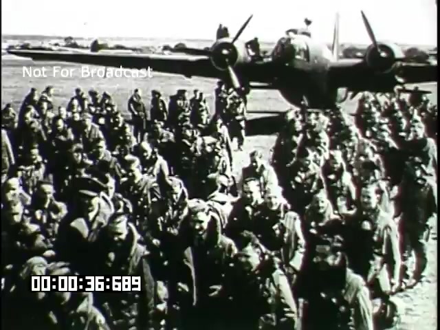 This 16mm still from the 'Battle of Britain' newsreel depicts a group of soldiers gathered on an airfield, with an aircraft visible in the background. The soldiers appear to be in military uniforms, possibly engaged in conversation or in a moment of camaraderie. The scene captures the atmosphere of wartime unity and reflects the historical context of the Battle of Britain. The image is monochromatic, typical of the era, conveying the gravity of the situation.