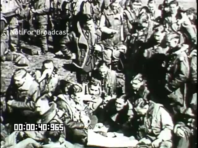 The still from the 'Battle of Britain' newsreel depicts a group of military personnel gathered closely together, likely pilots or aircrew. They are wearing flight jackets, showing signs of wear and fatigue. The atmosphere appears tense yet collaborative as they engage with one another, possibly discussing tactics or sharing experiences from combat. The scene captures the camaraderie and urgency of wartime, highlighting the human aspect of the conflict. The film is likely black and white, reflecting the era and medium.