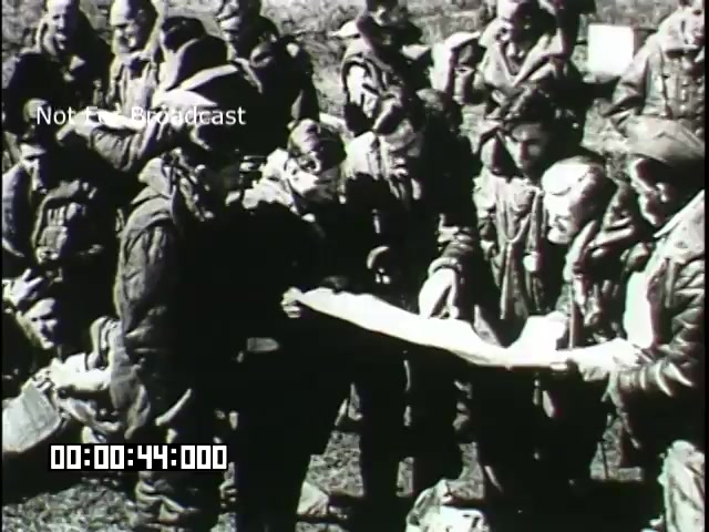 The still from the 'Battle of Britain' newsreel depicts a group of military personnel gathered around a map, engaged in discussion. They appear focused and serious, highlighting the tension and urgency of the situation during the battle. The image captures a moment of collaboration and strategy among the airmen, showcasing their determination amidst the conflict. The overall tone evokes a sense of camaraderie and the gravity of their mission.
