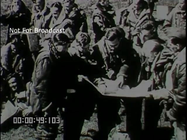 The still from the 'Battle of Britain newsreel' depicts a group of military personnel, likely pilots, gathered around a map. They are dressed in flight gear and appear to be engaged in a discussion, possibly planning their next mission. The setting suggests a sense of urgency and teamwork, characteristic of the strategic operations during the Battle of Britain. The image is in black and white, reflecting the historical context of the 16mm film format.