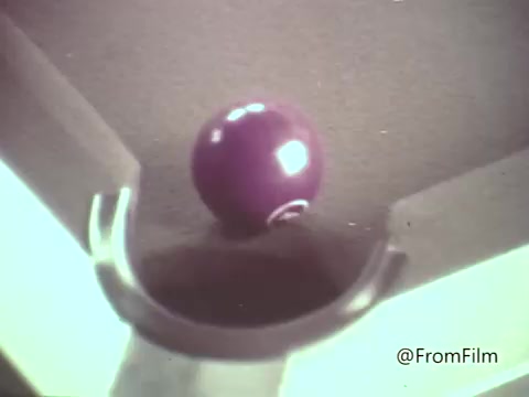 The still features a purple sphere, resembling an M&M's candy, rolling toward a hole on a surface that resembles a pool table. This imagery plays into the whimsical and playful nature typical of 1960s animated commercials for M&M's candies, where candies are often anthropomorphized and engaged in humorous scenarios. The overall aesthetic captures the vibrant color scheme and animation style of that era, appealing to a youthful audience.