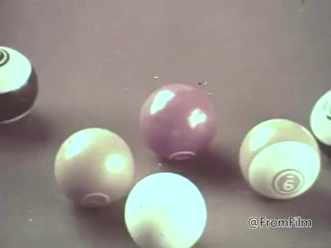 The still features a close-up of several billiard balls arranged on a surface, resembling a playful scene. The colors, including shades of purple and light blue, evoke a light-hearted, animated aesthetic typical of 1960s commercials. This aligns with the whimsical style usually seen in animated M&M's candy advertisements from that era, blending fun and vibrancy to attract viewer attention.