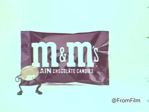 The still features a whimsical animated representation of an M&M's candy character standing next to a purple M&M's package. The character has limbs and a cheerful expression, embodying the playful spirit of the 1960s commercials. The overall aesthetic reflects the vibrant and fun style typical of that era's advertising, aimed at capturing the attention of viewers with its lively animation and colorful branding.