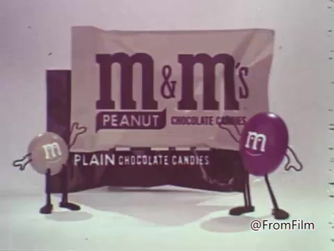 The still features two anthropomorphized M&M's candies in an animated style typical of the 1960s. One candy is brown and represents plain chocolate, while the other is purple, representing peanut chocolate. They stand beside a prominently displayed packaging of both types of M&M's, which is styled with the brand logo and text indicating 'PEANUT' and 'PLAIN.' The characters have arms and are animatedly posing, embodying a playful and friendly vibe characteristic of the era's advertising.
